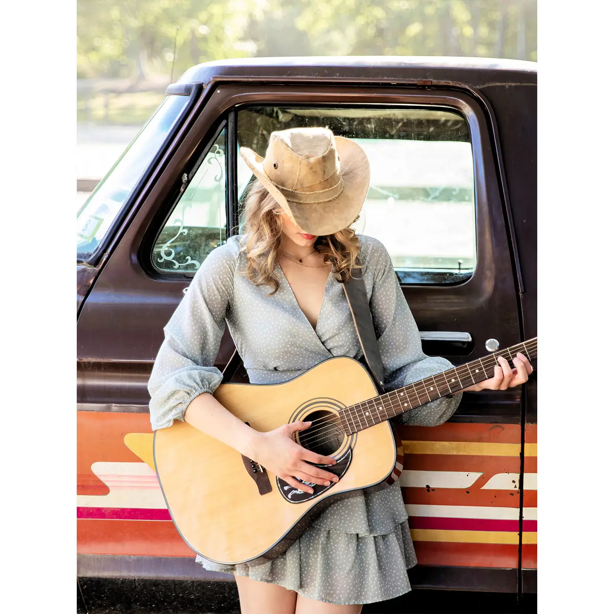 Model standing in front of a truck wearing the guitar strap over her shoulder with an acoustic guitar.